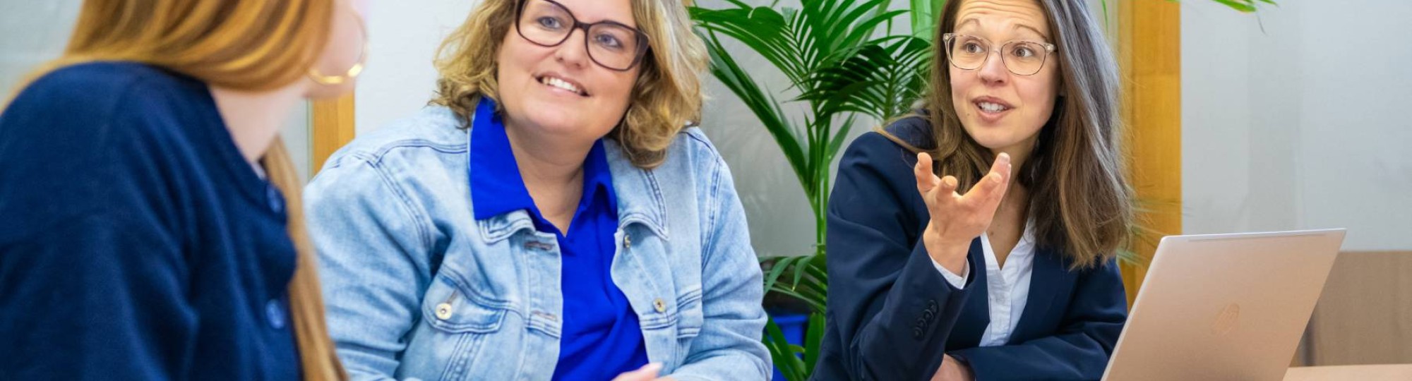 Drie mensen aan tafel in gesprek met elkaar, met een hoop papieren op tafel en een laptop
