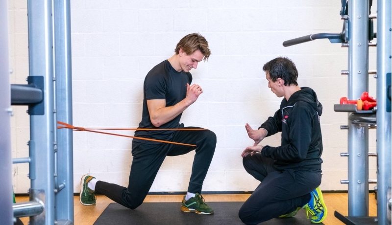 Student en docent oefenen in gymzaal, jongen zit op 1 knie en stretcht zijn been.
