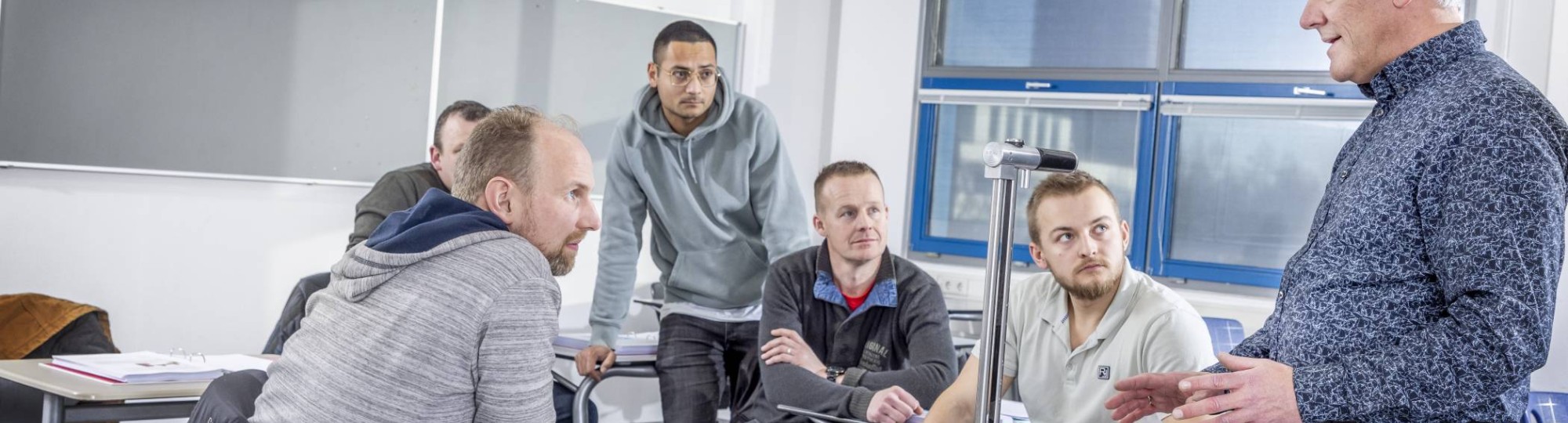 5 mannen zittend en staand aan een tafel luisteren aandachtig naar de docent