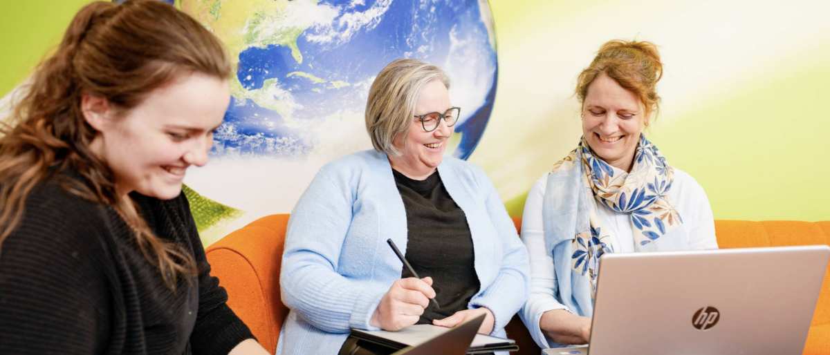 Vrouwen lachen op oranje bank, 1 vrouw maakt aantekeningen. Wereldbol op achtergrond.
