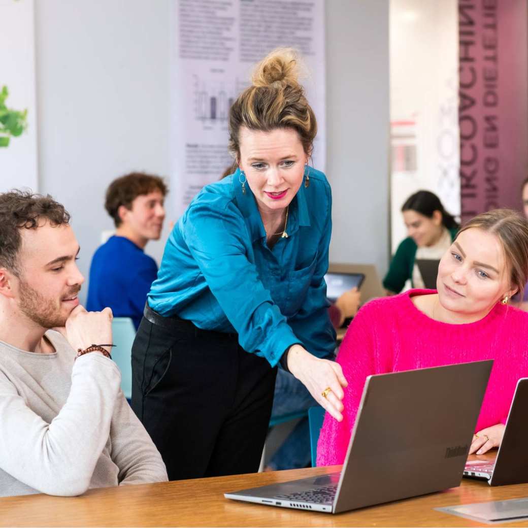 Docent helpt studenten door iets aan te wijzen op een laptop die voor de student op tafel staat. 