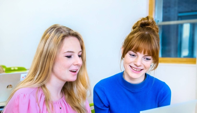 Twee vrouwelijke studenten leraar Geschiedenis zitten naast elkaar en kijken lachend naar het scherm (laptop) dat voor hun staat.