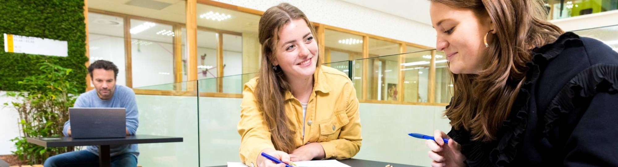 ALPO-student zit samen met haar docent te sparren aan een tafeltje in de gang van het I/O gebouw.