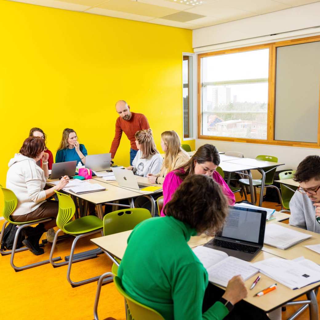 De docent loopt rond in het klaslokaal. De studenten zijn aan het werk. 