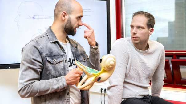 Man wijst zijn neus aan, terwijl hij een anatomiemodel van een oor vasthoudt en naar een andere man kijkt.