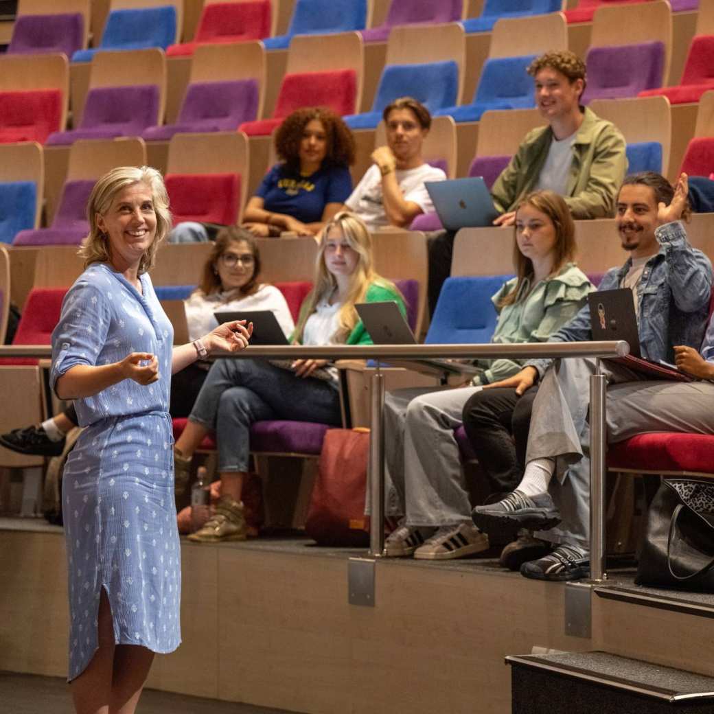 Docente kijkt lachend in de camera en geeft hoorcollege in collegezaal. 