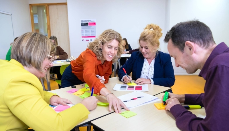 Een groepje van vier mensen in gesprek met post-its op tafel in een lokaal met anderen