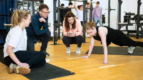 Student staat in een plank positie, 3 studenten kijken toe. 
