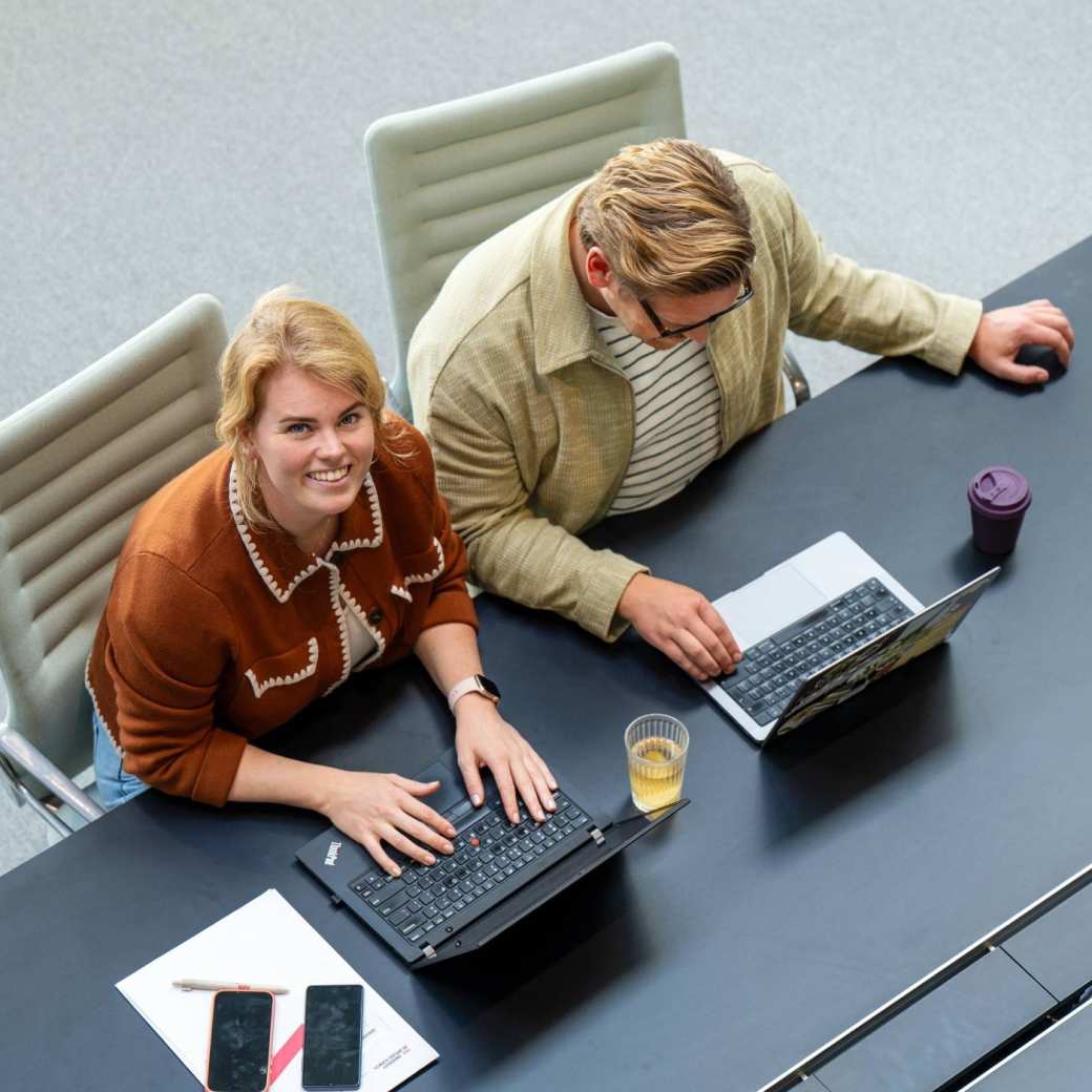 Twee studenten zitten te werken achter laptop en kijken lachend in camera
