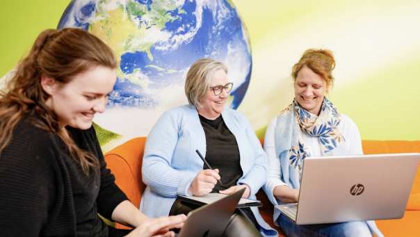 Vrouwen lachen op oranje bank, 1 vrouw maakt aantekeningen. Wereldbol op achtergrond.