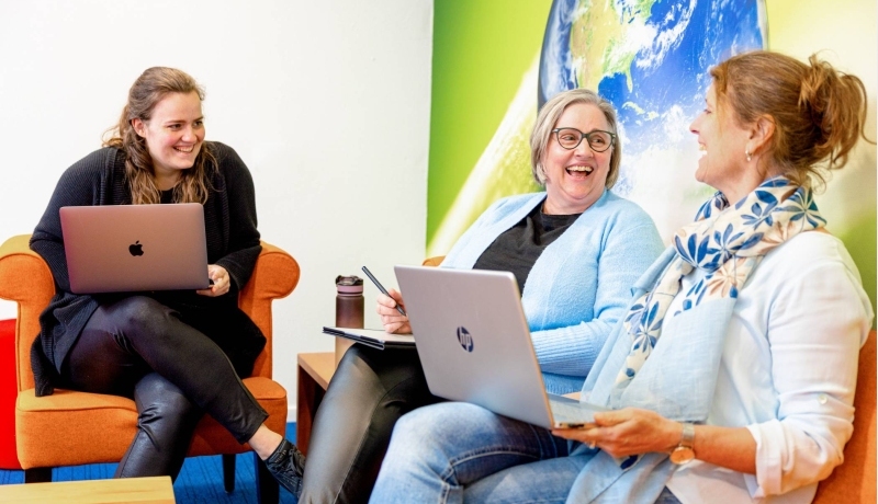 3 vrouwen lachen. Oranje banken en een wereldbol op de achtergrond.