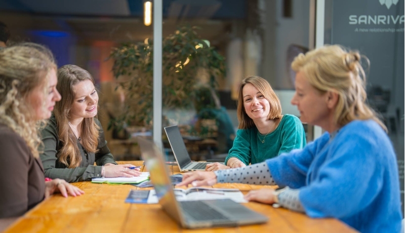 3 studenten en één docent van de master Duurzaamheidstransities zitten aan een tafel en overleggen. Eén student kijkt in de camera en lacht.