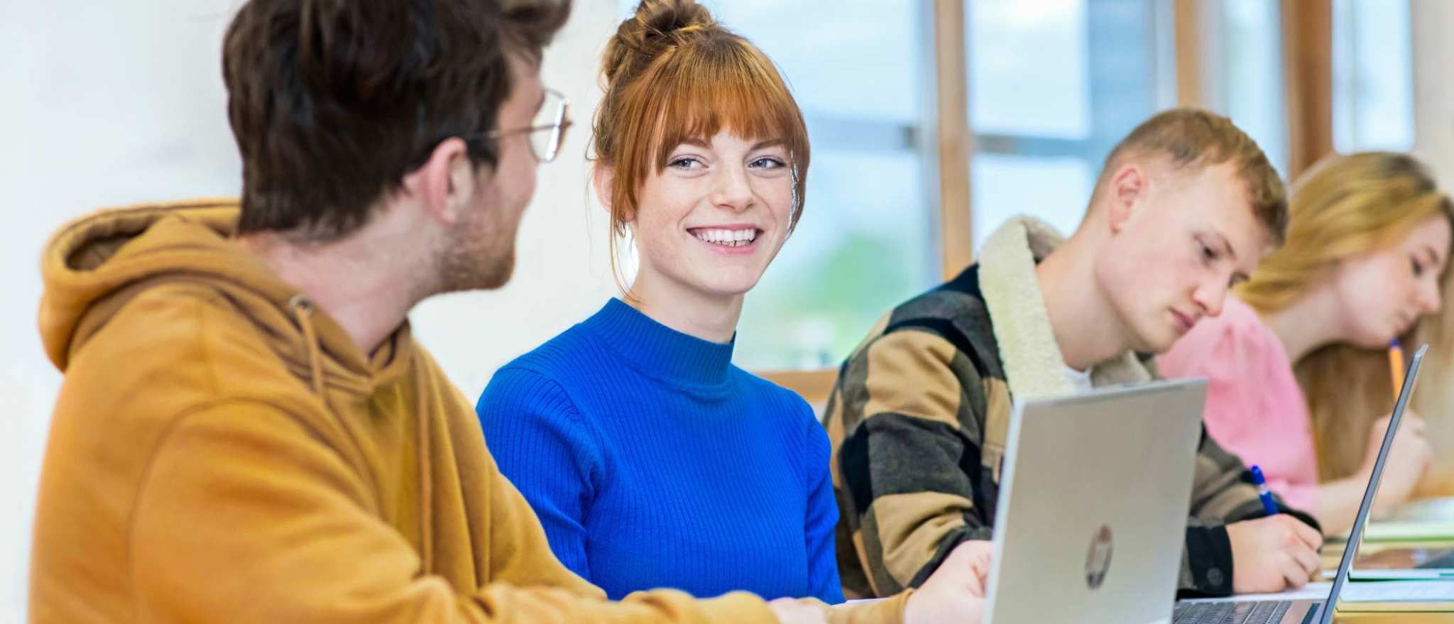 Vier studenten zitten op een rij. Een student specifiek is scherp in beeld en ze lacht naar de persoon naast haar.