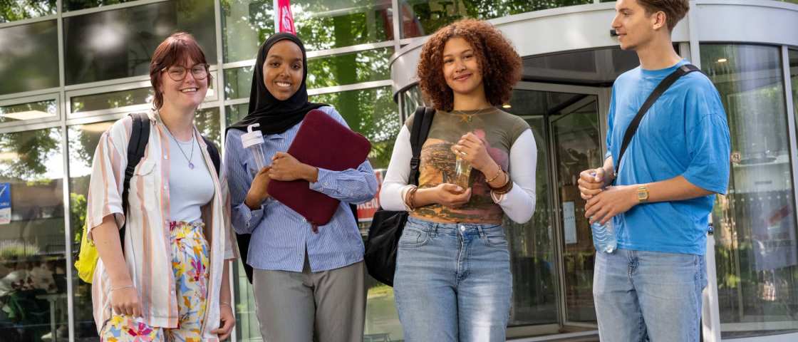 Vier studenten staan bij gebouw LvS10 bij draaideur, buiten. Op een rijtje. 