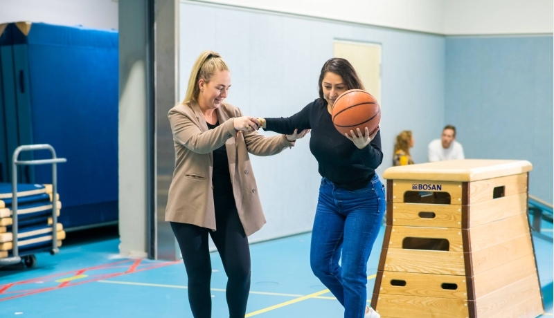 Twee studenten waarbij één student op een skateboard staat met een bal in de hand en de andere student haar begeleidt
