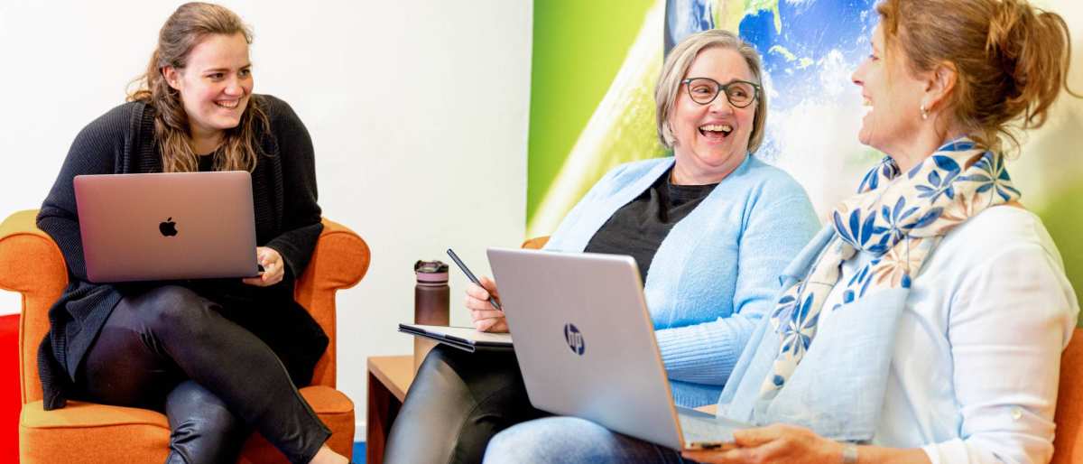 3 vrouwen lachen. Oranje banken en een wereldbol op de achtergrond.