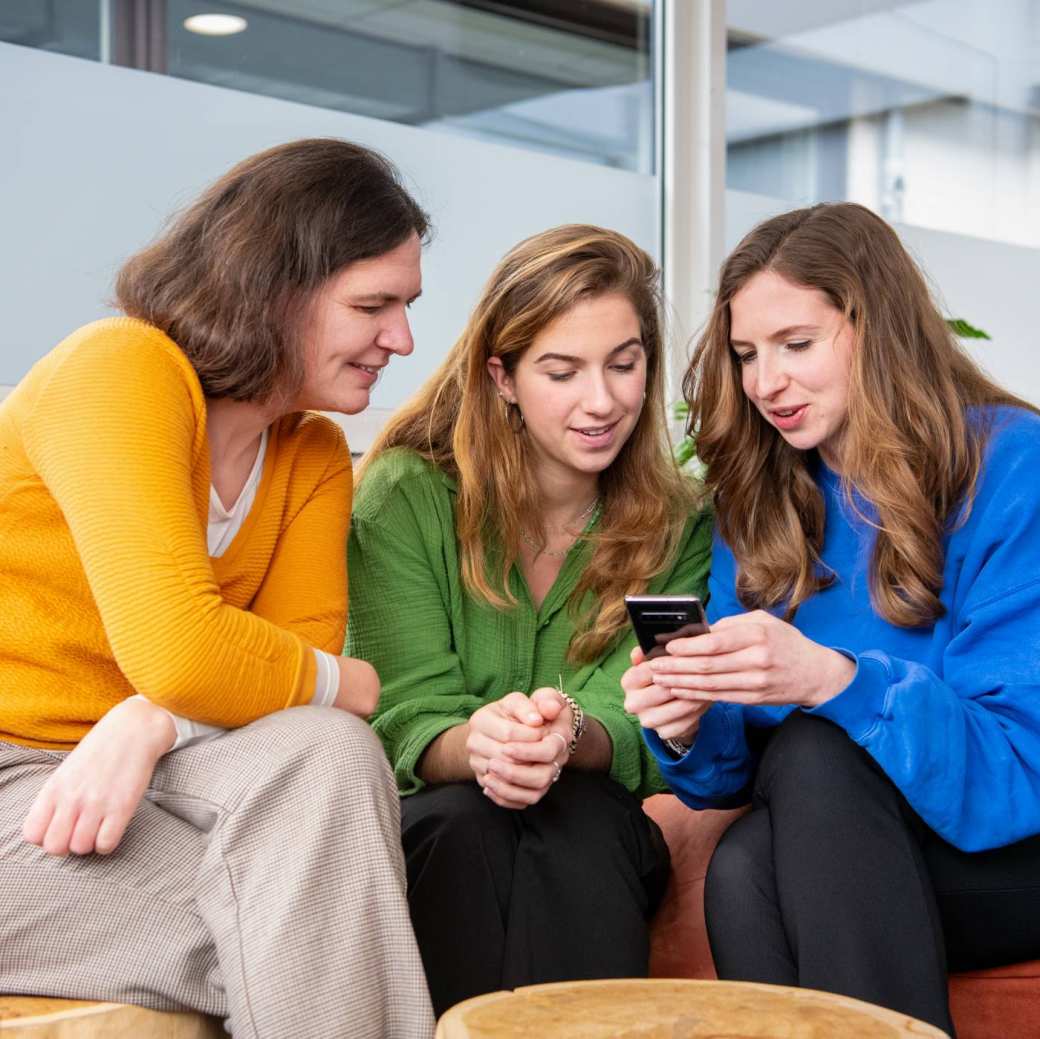 3 Studenten zitten samen op krukjes en kijken op een telefoon.