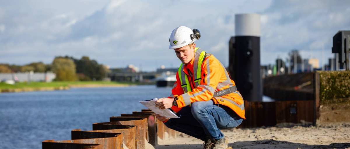 Man met veiligheidshelm en oranje werkjas zit gehurkt bij een damwand aan een kanaal.