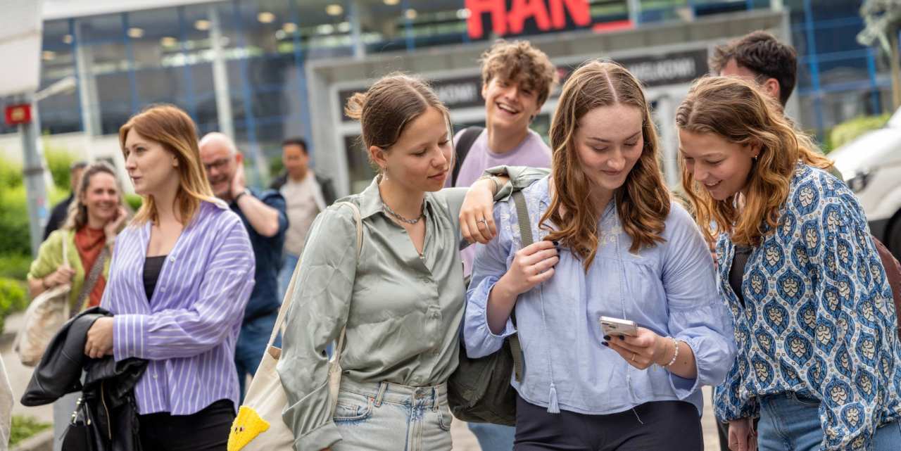 Studenten staan met een groepje buiten bij de ingang van gebouw R26, kijken op een telefoon. 