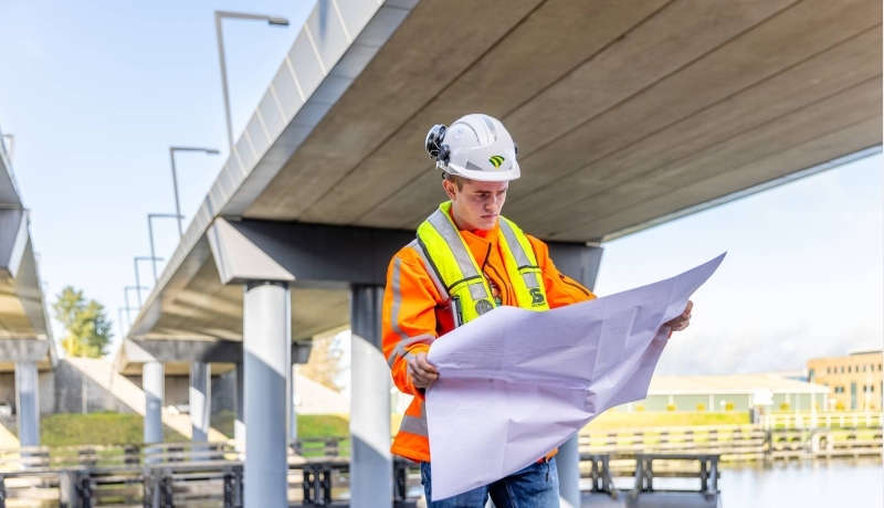 Man met veiligheidshelm en oranje werkjas checkt de bouwtekening onder een viaduct in aanbouw.