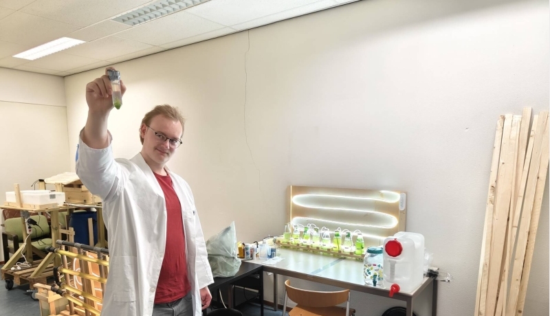 Bas Emaus, blond haar, brilletje op, in een labjas met zijn algenparels in zijn hand en achter hem zijn installatie