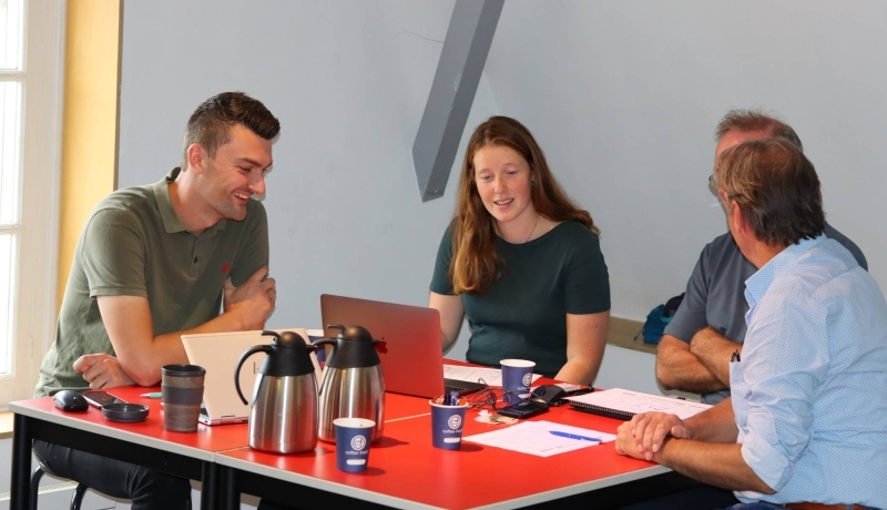 Een groepje van drie mensen overlegt en tekent aan een rode tafel. Laptops, koffiekannen en bekertjes in beeld. De foto is gemaakt tijdens de opstart van de deeltijdmaster Circulaire Economie in de educatieruimte in het Nederlands Watermuseum. 9 September 2022