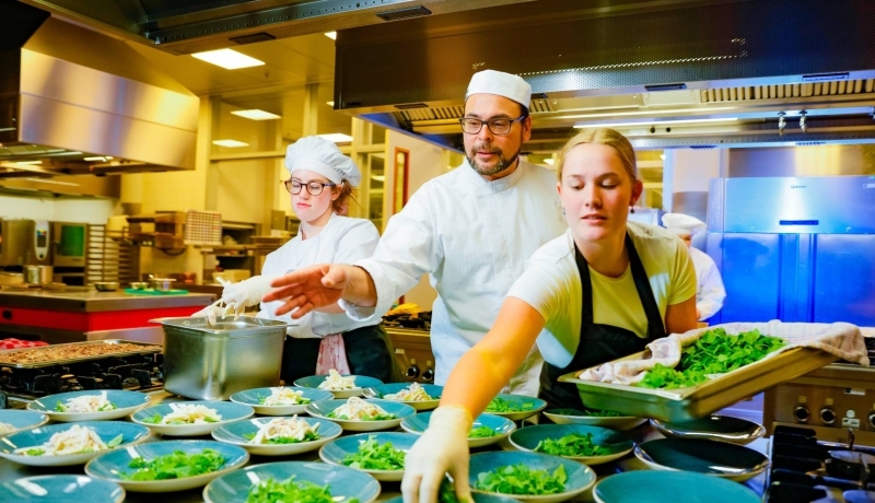 Student legt sla op een bord terwijl de chefkok naast haar instructies geeft.