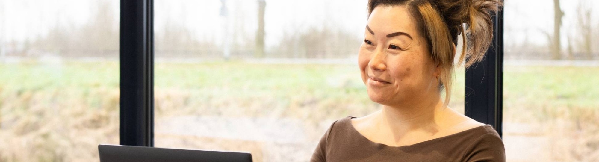 Dame poseert met een glimlach, achter een laptop. Een glas water staat naast haar op het bureau.
