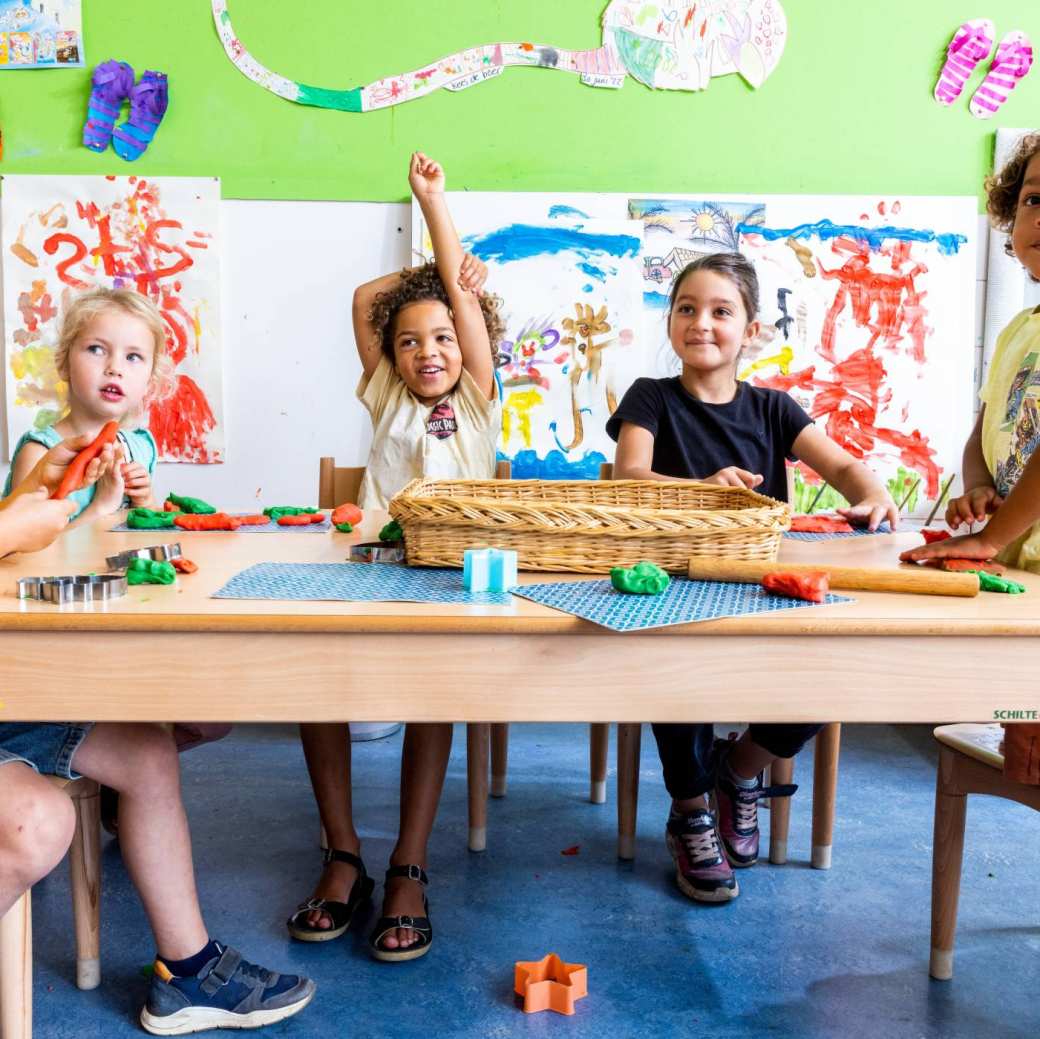Vijf kinderen zitten aan hun tafel in de klas, waarvan één kind zijn vinger opsteekt om een vraag te stellen