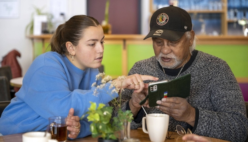 Een oudere man met een Ipad, een jonge dame wijst hem hier iets op aan.