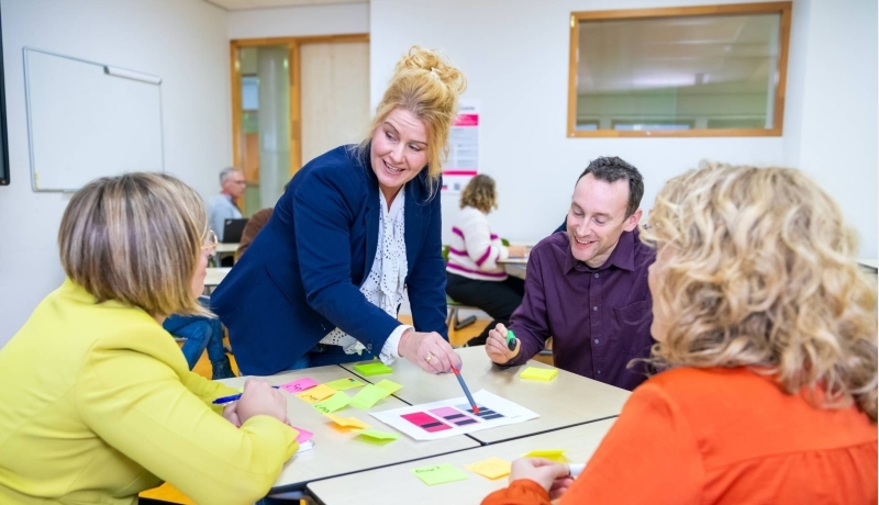Een groepje van vier mensen in gesprek met post-its op tafel in een lokaal met anderen