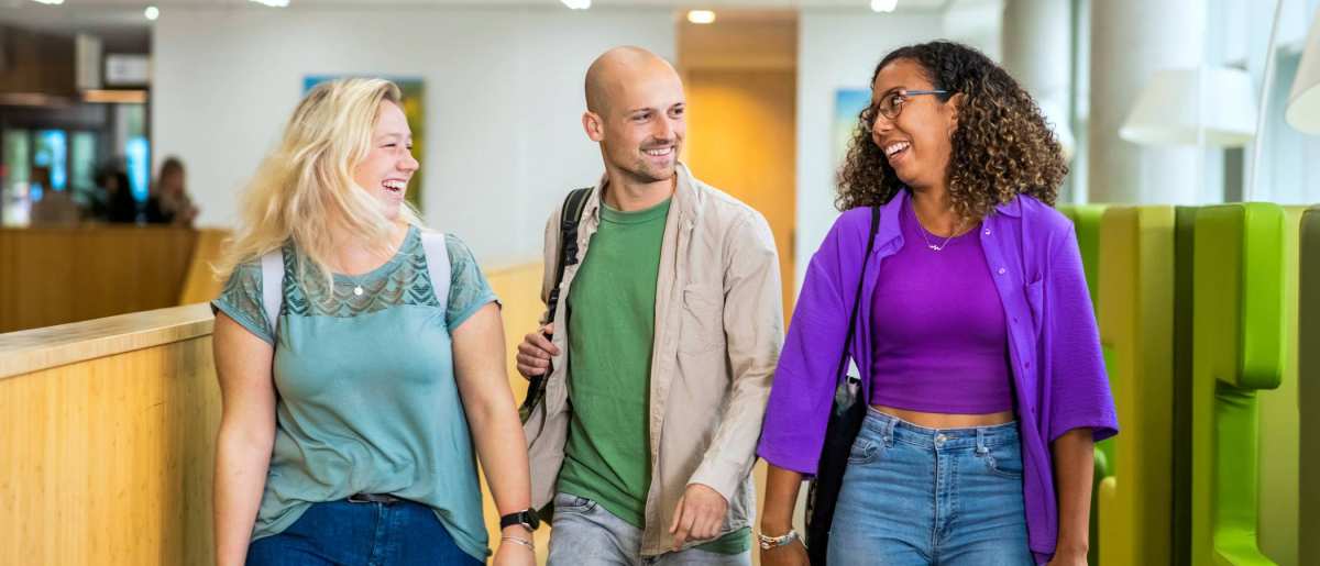 Drie studenten Leraar Wiskunde lopen lachend naast elkaar op de gang in de school

