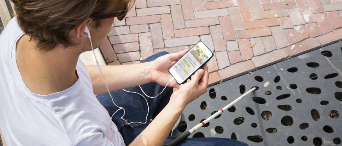 Jongen zit op een bankje en is met zijn mobiele telefoon bezig. Voor hem ligt zijn taststok.