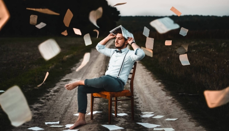man met wit overhemd, vlinderdas en bretels zit op een stoel, op een zandweg. Hi houdt een boek boven zijn hoofd en losse pagina's vliegen in het rond