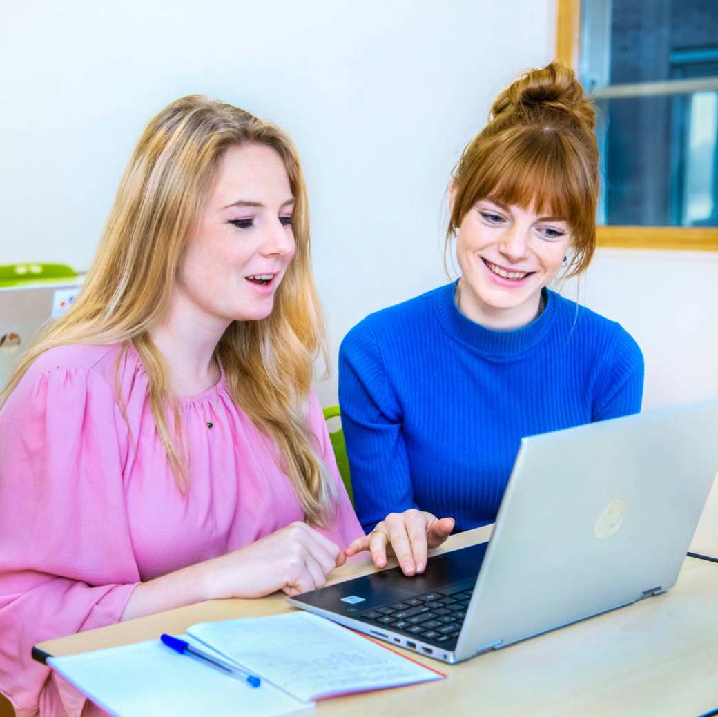 Twee vrouwelijke studenten leraar Geschiedenis zitten naast elkaar en kijken lachend naar het scherm (laptop) dat voor hun staat.