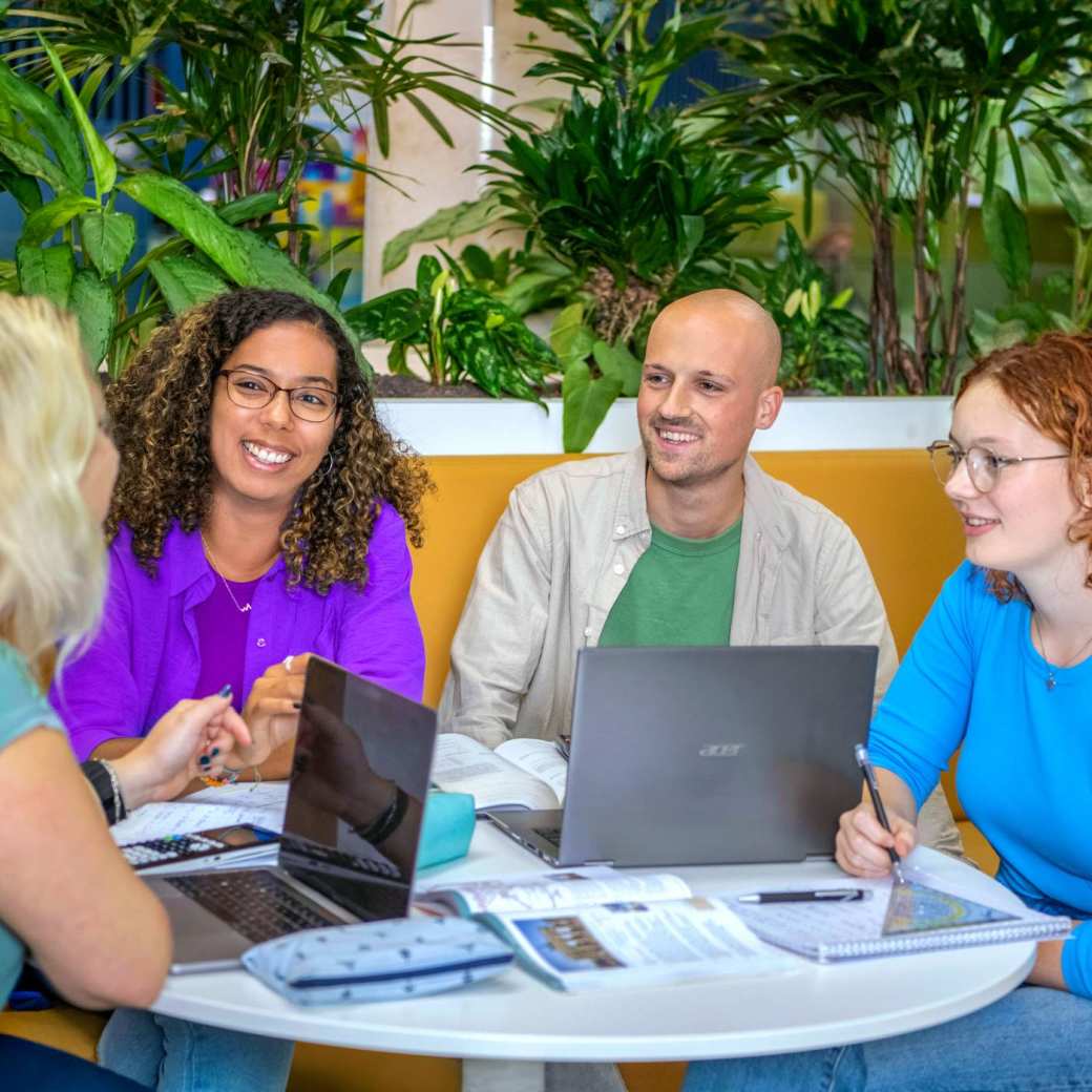 Vier studenten Leraar Wiskunde zitten aan een ronde tafel te werken. Er liggen laptops, etuis en schriften op de tafel.