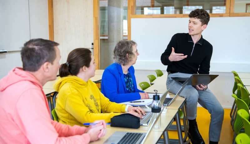 Foto Master Academie Educatie. 4 masterstudenten zitten in een lokaal. 1 van hen zit op een bureau en legt met een laptop in z'n hand wat uit aan de rest.