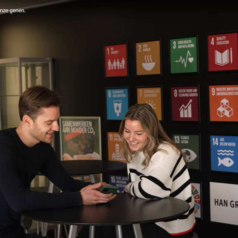 Still van de wand van de Green Office met alle SDG's en twee studenten van de GO ervoor.