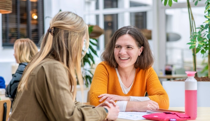 Vrouw in oranje trui lacht naar medestudent.