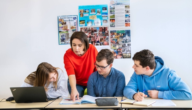 Drie studenten zitten op een rij en de docent staat achter hen. Ze wijst in het boek en de studenten kijken mee.