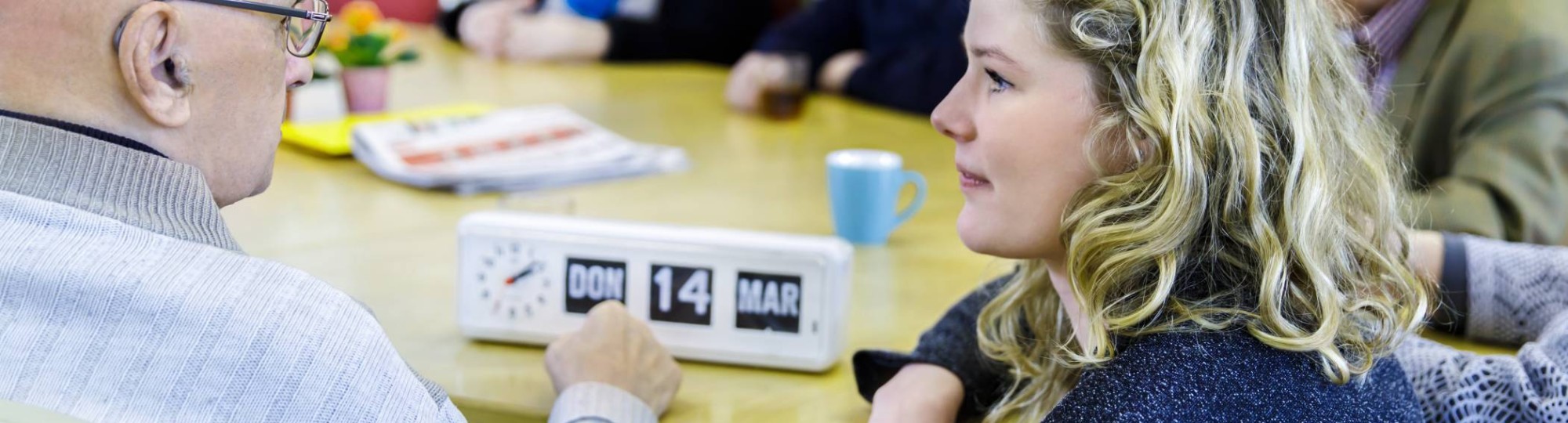 bijeenkomst wijkatelier lindenholt- aan tafel studente hurkt en geeft uitleg kalenderklok