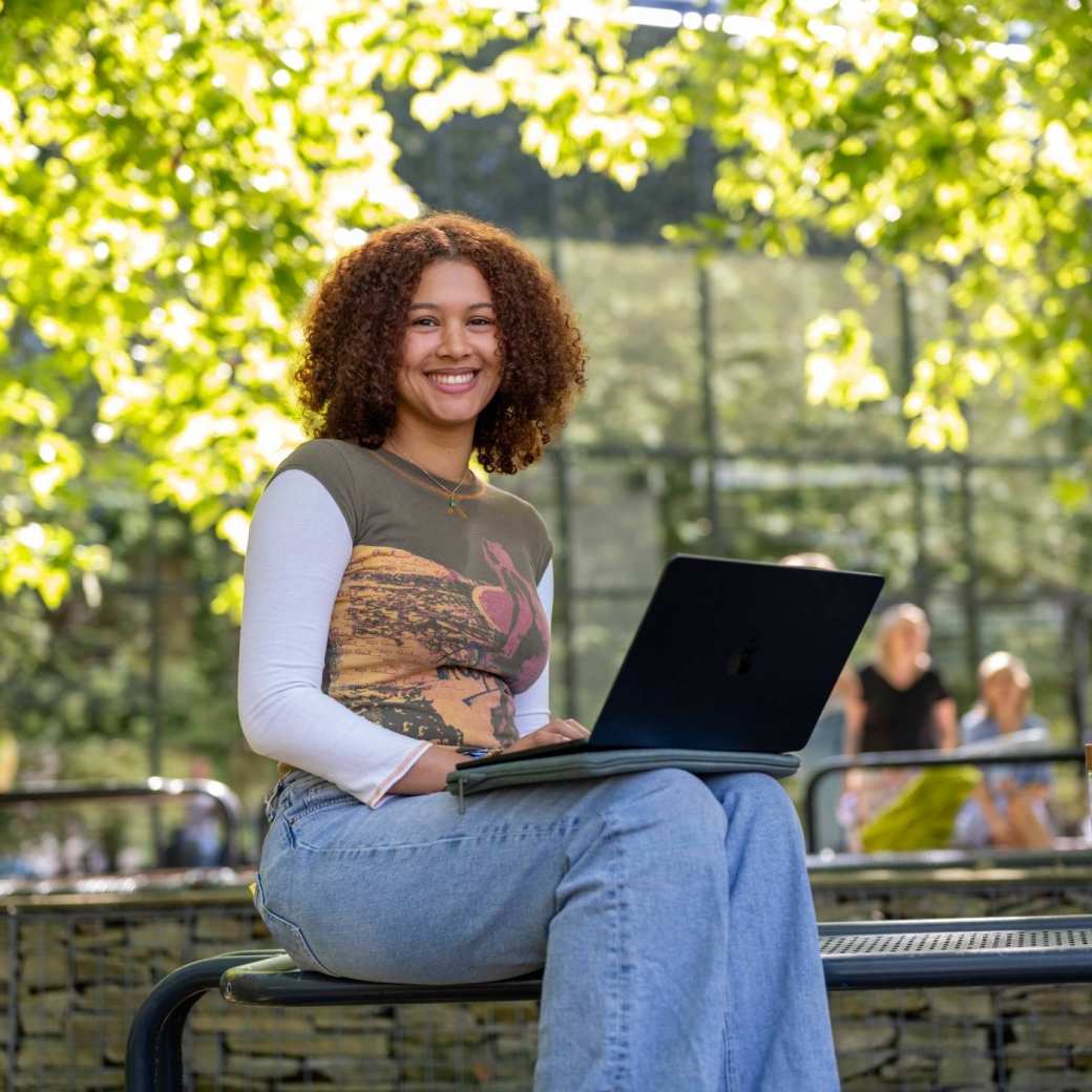 Studente kijkt in de camera, met laptop op schoot, zittend op de tafel, buiten bij K33.