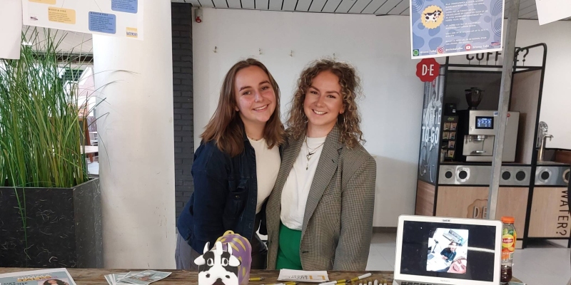 Silke Vondel en Marhot Ausems presenteren de opbrengsten van hun onderzoek op het PZW evenement. Links een vrouw met lichtbruin sluik haar en rechts blonde krullen en een grijze blazer. Ze staan in kraam volgehangen met posters.
