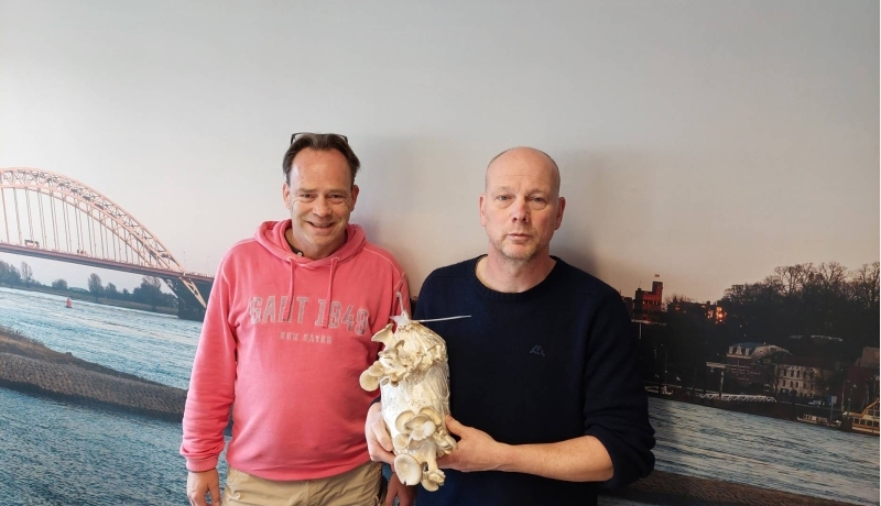 Mark Heussen en René Klep poserend voor een fotomuur met Nijmegen aan de Waal erop. Rene met oesterzwammen groeiend op een voedingsbodem van onder andere koffiedik.