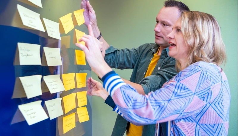 blonde vrouw met blauw en roze vest en man kort haar en groene blouse met geel tshirt, hangen gele post-its op een glazen wand