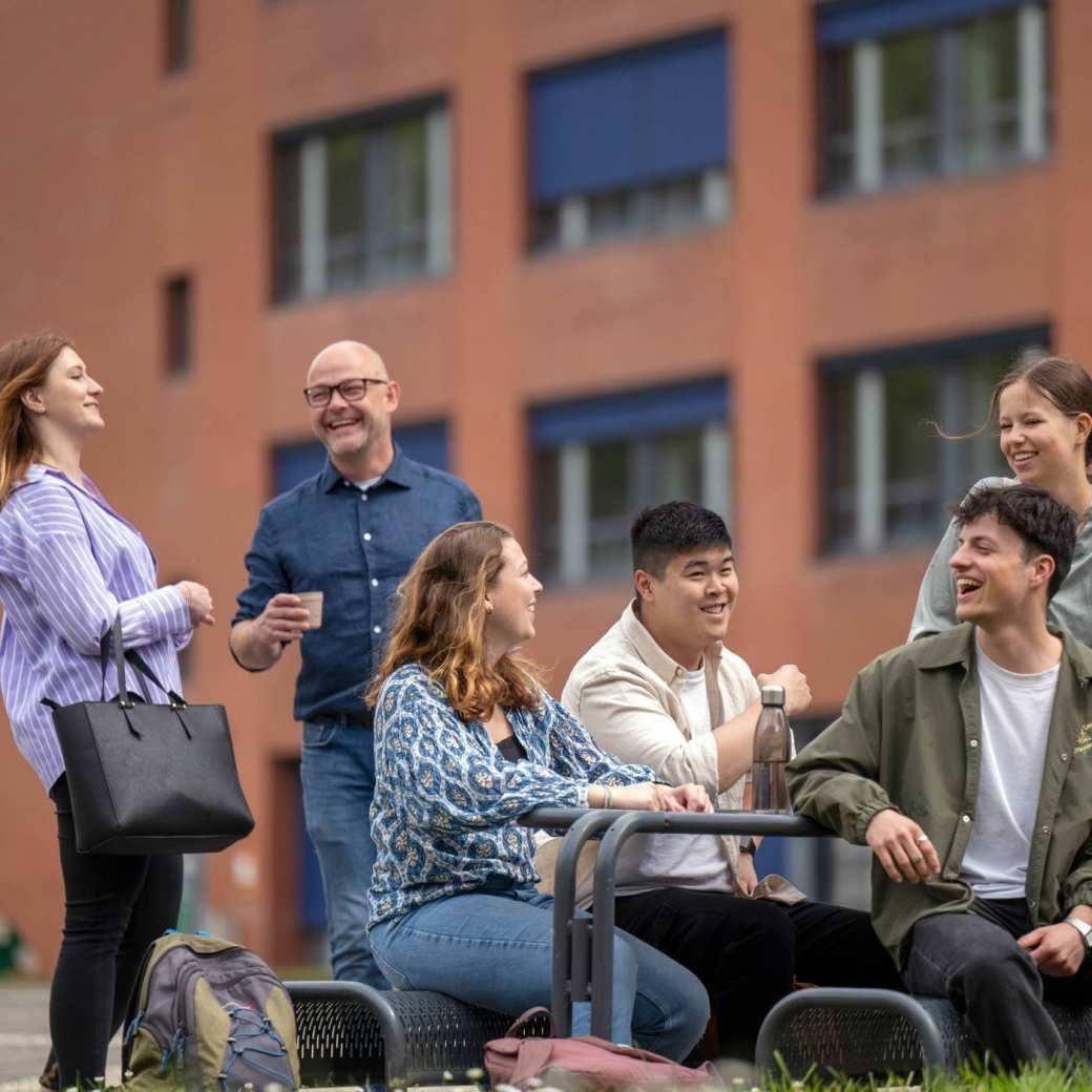 Studenten zitten buiten aan picknicktafel, hebben plezier en lachen. Op campusterrein Arnhem. 