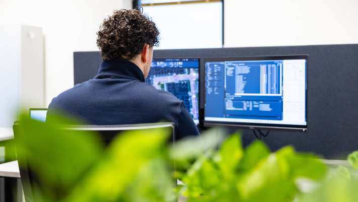 Man werkt op twee beeldschermen aan een bouwontwerp. Op de voorgrond staat een plant