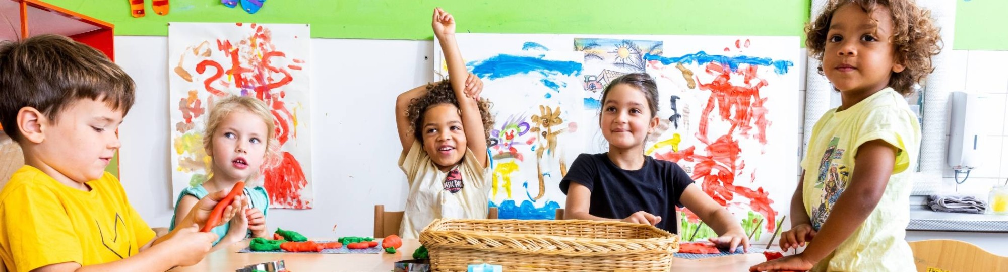 Vijf kinderen zitten aan hun tafel in de klas, waarvan één kind zijn vinger opsteekt om een vraag te stellen. Foto voor de opleiding leraar basisonderwijs