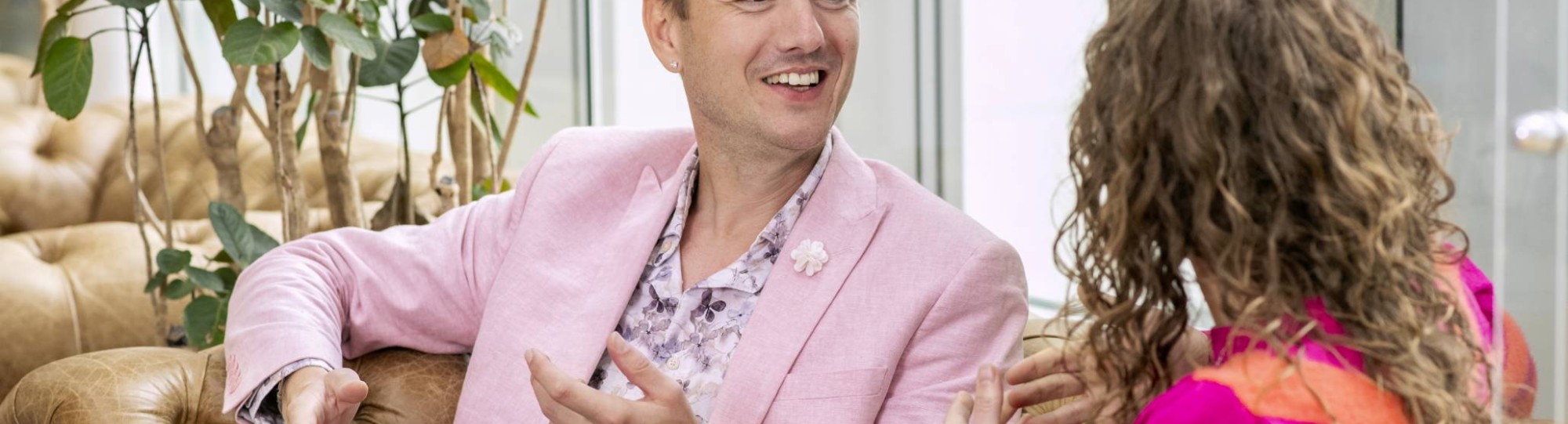 Mannelijke student met roze/paarse  jasje lacht naar zijn medestudent die naast hem op de sofa zit. 