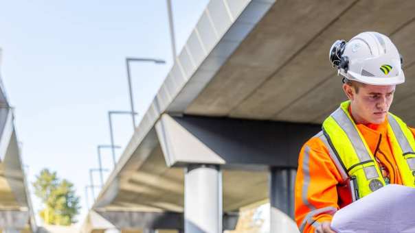 Man met veiligheidshelm en oranje werkjas checkt de bouwtekening onder een viaduct in aanbouw.